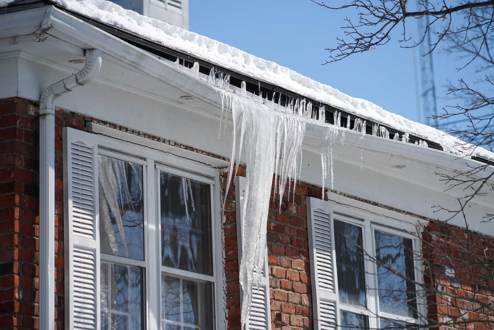 De longues stalactites de glace pendent du toit enneigé d'une maison en briques rouges, sous un ciel d'un bleu éclatant.
