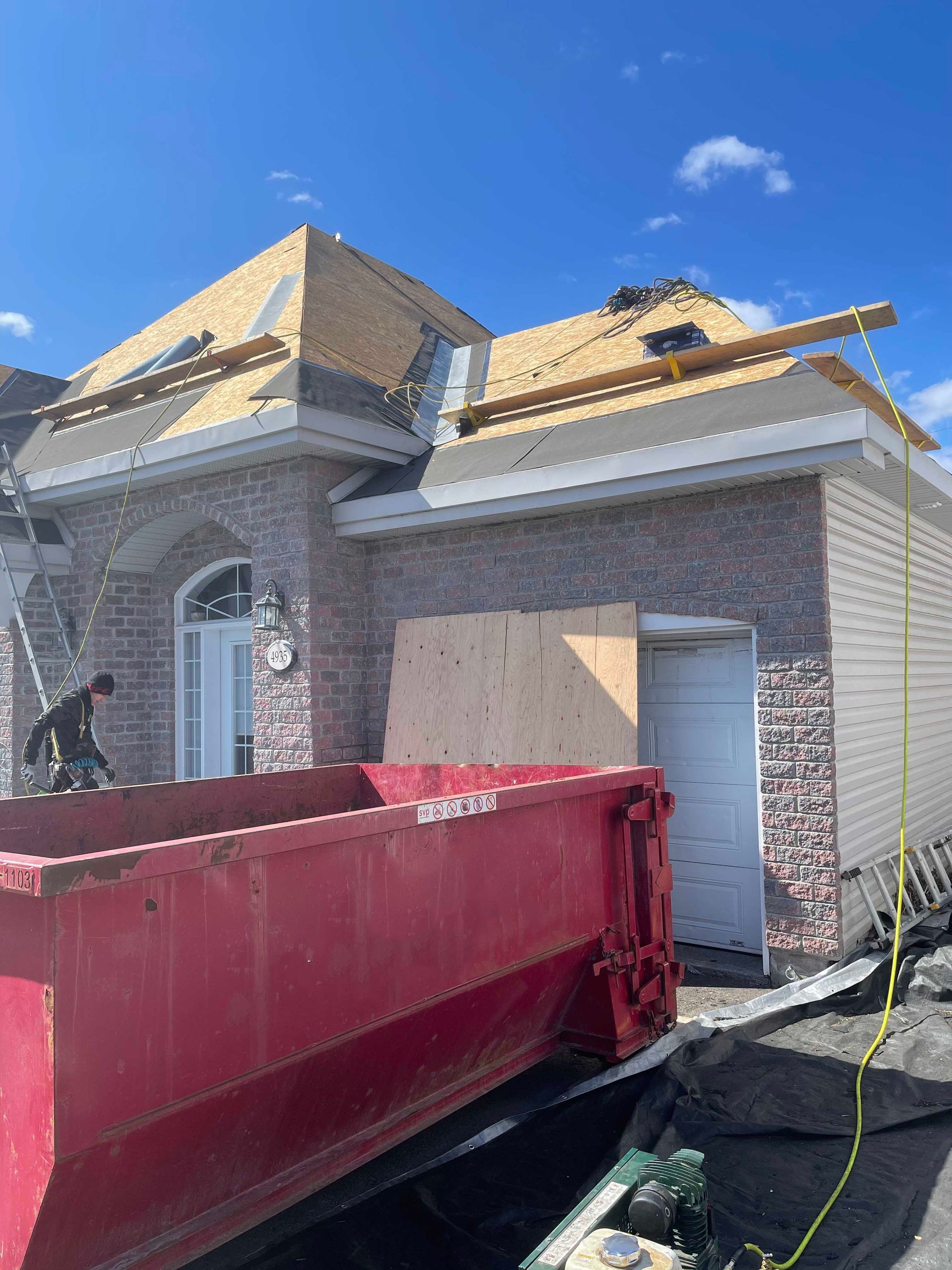 Une maison en briques en construction, avec une porte recouverte de contreplaqué, une benne à ordures rouge au premier plan et des échafaudages.