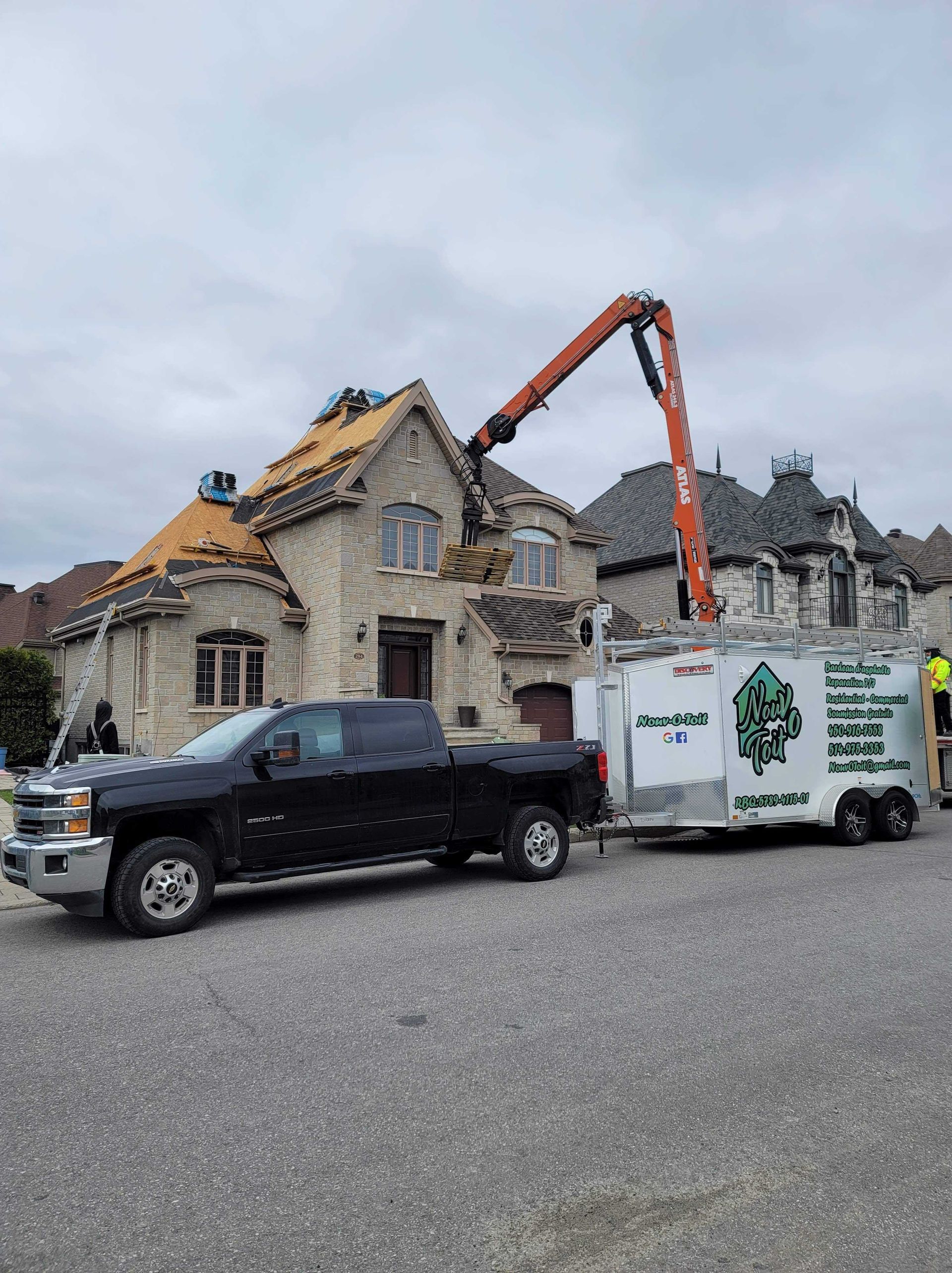 Un pick-up noir avec une remorque blanche est garé devant une maison en construction avec une grue sur le toit.