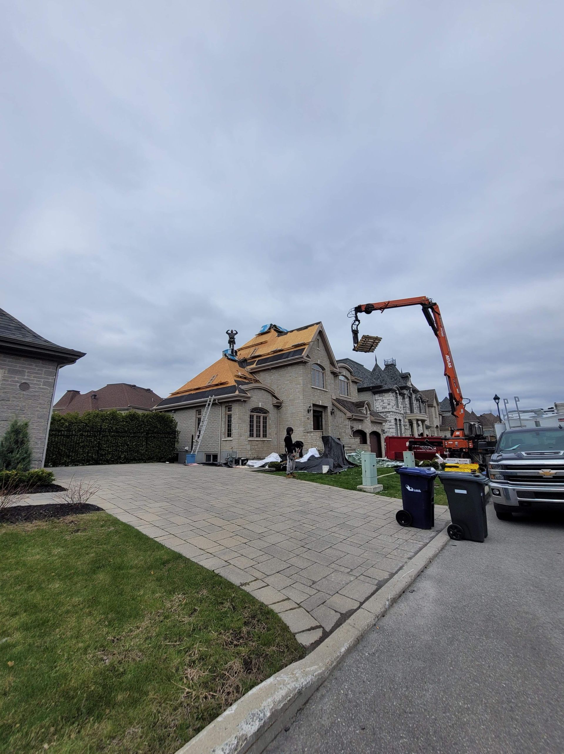 Une maison en pierre en construction, la toiture apparente, est desservie par une grande grue orange dans une rue résidentielle.