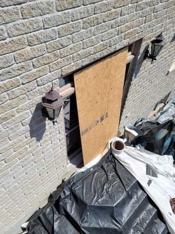 Un panneau de contreplaqué recouvre une grande ouverture dans un mur d'un bâtiment en briques beiges, avec des bâches de chantier en dessous.