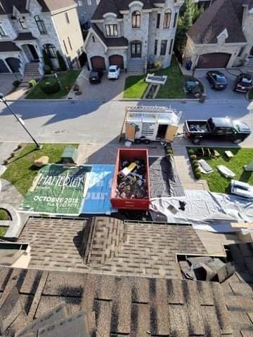 Vue en plongée d'un remplacement de toiture résidentielle, montrant un conteneur à déchets rouge et des bâches de protection dans l'allée en contrebas.