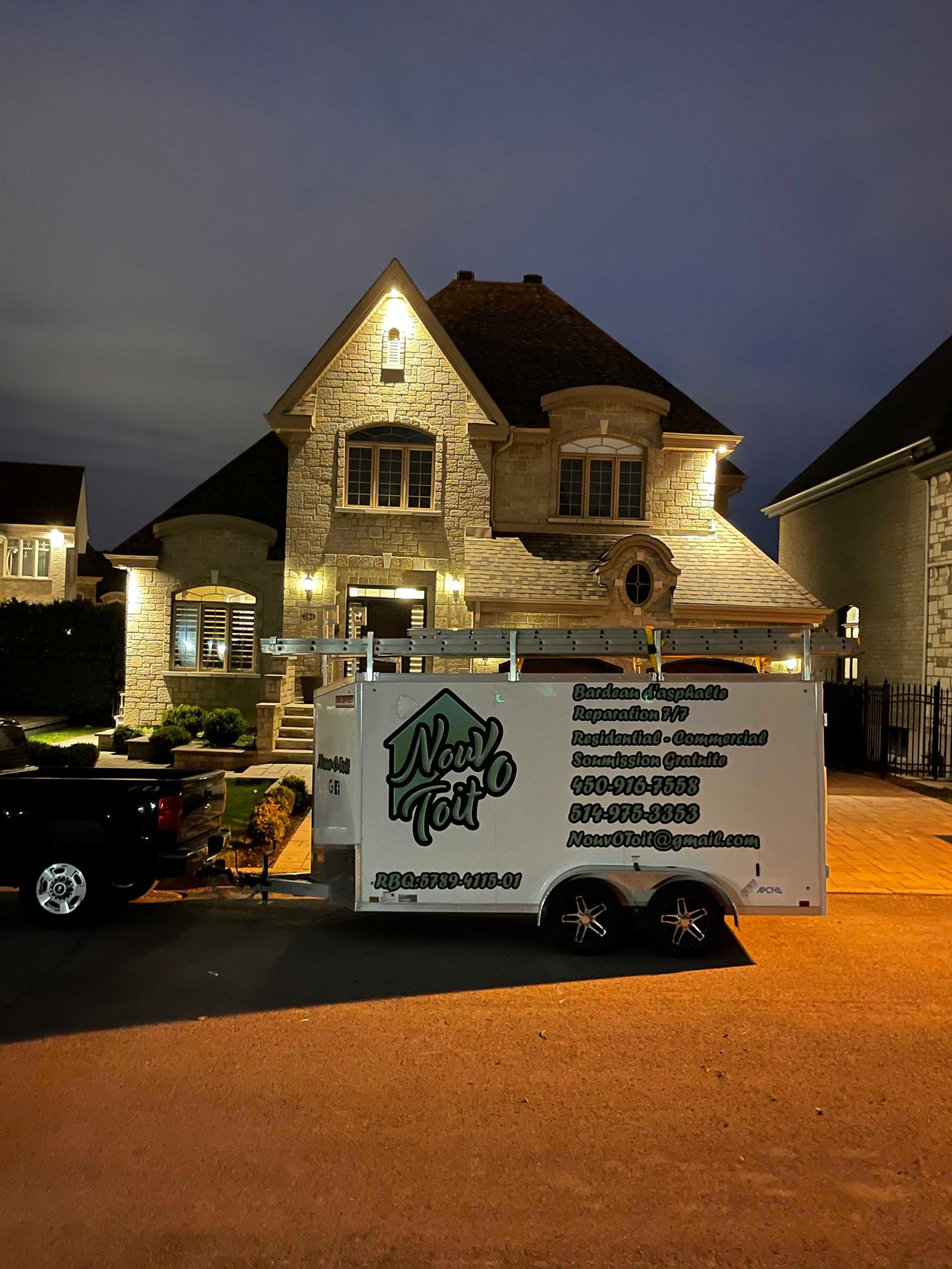 Une remorque de chantier arborant la marque « New Age » est garée devant une maison en pierre au crépuscule, éclairée par des projecteurs extérieurs.