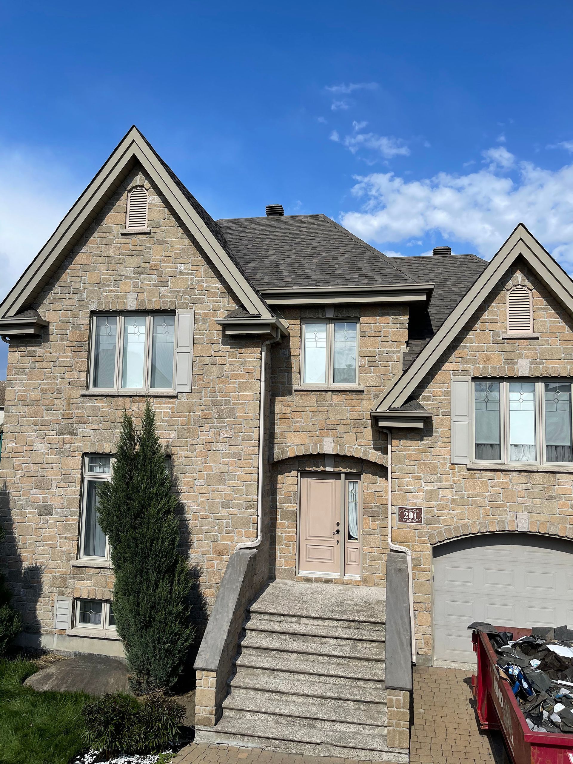 Une maison en pierre beige de deux étages avec un toit pentu, des pignons, un escalier d'entrée et une benne à gravats rouge dans l'allée.