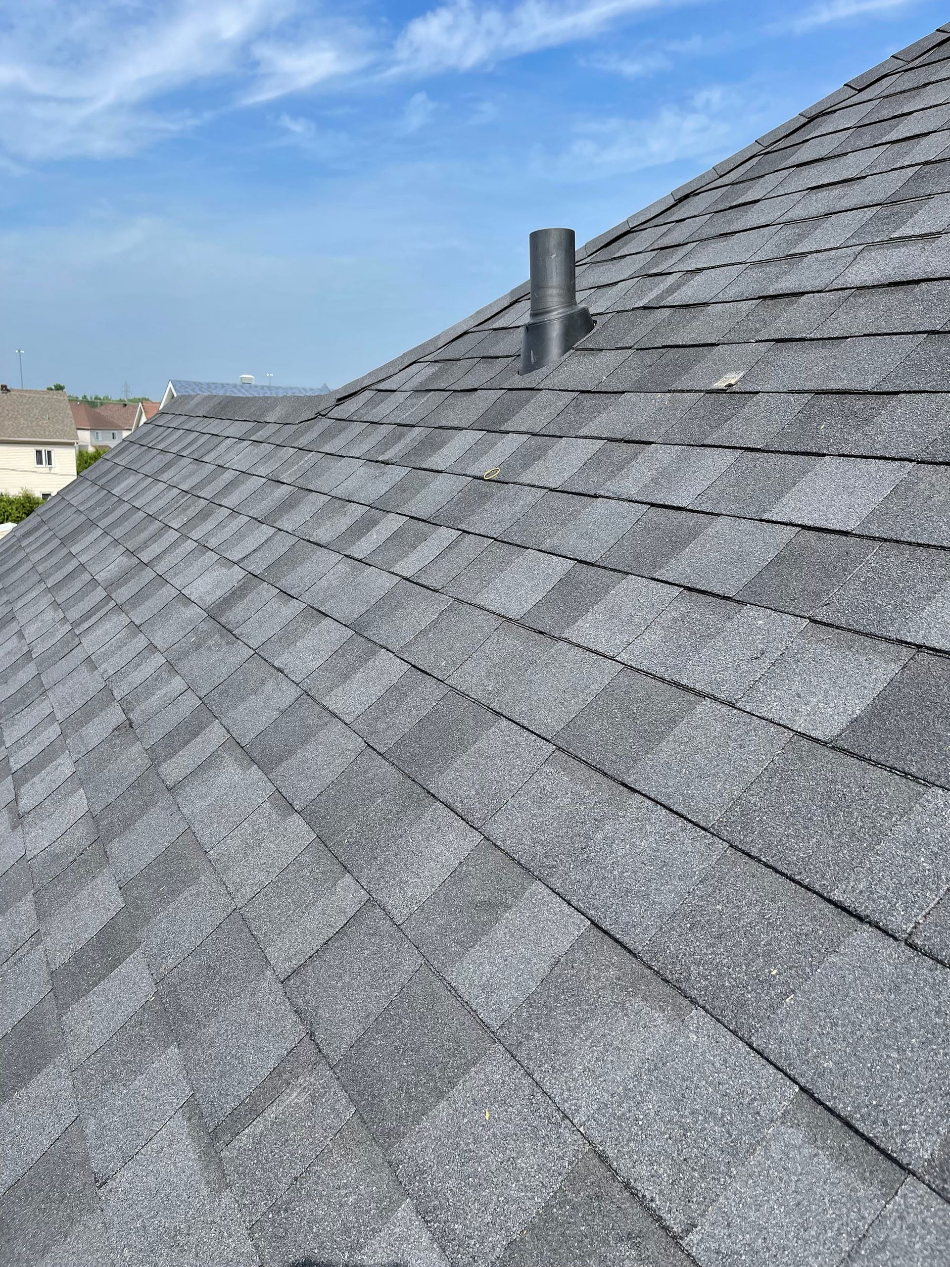 Un toit en bardeaux d'asphalte gris sous un ciel d'un bleu éclatant, d'où part un unique tuyau d'évacuation noir qui s'élève vers le ciel.