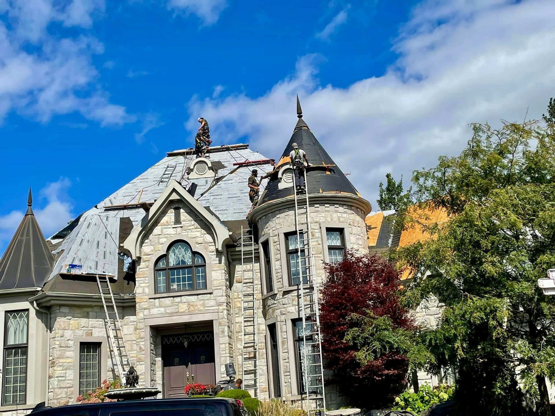 Des ouvriers réparent le toit d'une grande maison en pierre avec une tourelle, sous un ciel bleu.
