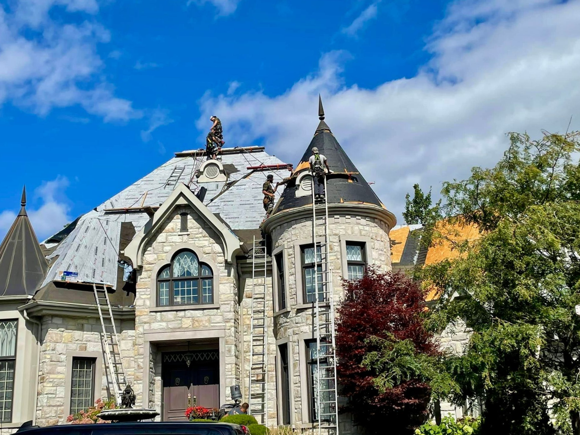 Des ouvriers, juchés sur des échelles, réparent le toit d'une grande maison en pierre dotée d'une tourelle conique proéminente, sous un ciel d'un bleu éclatant.