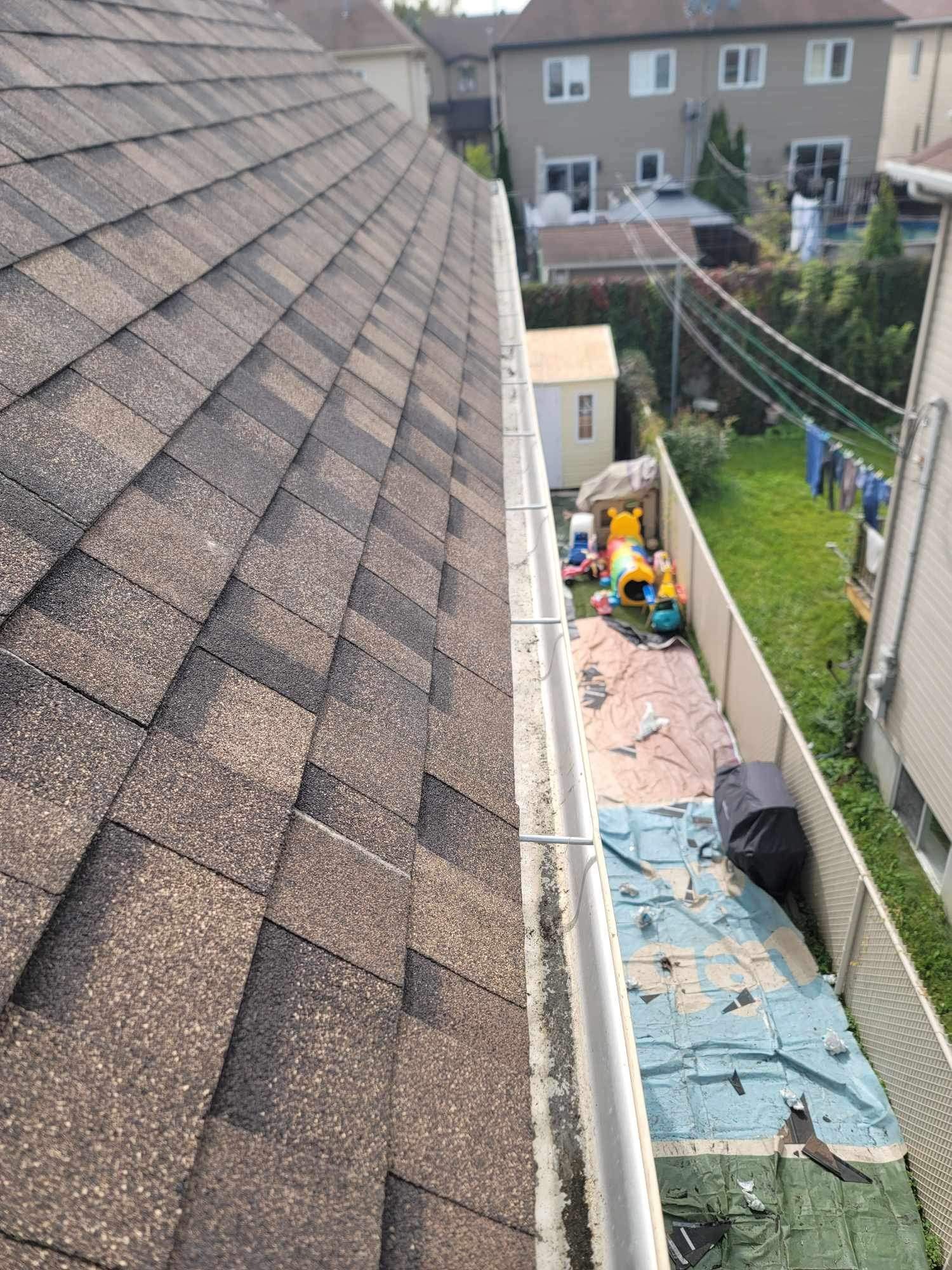 Vue en plongée depuis une gouttière longeant le bord d'un toit en bardeaux d'asphalte, en direction d'une cour arrière et des maisons voisines.