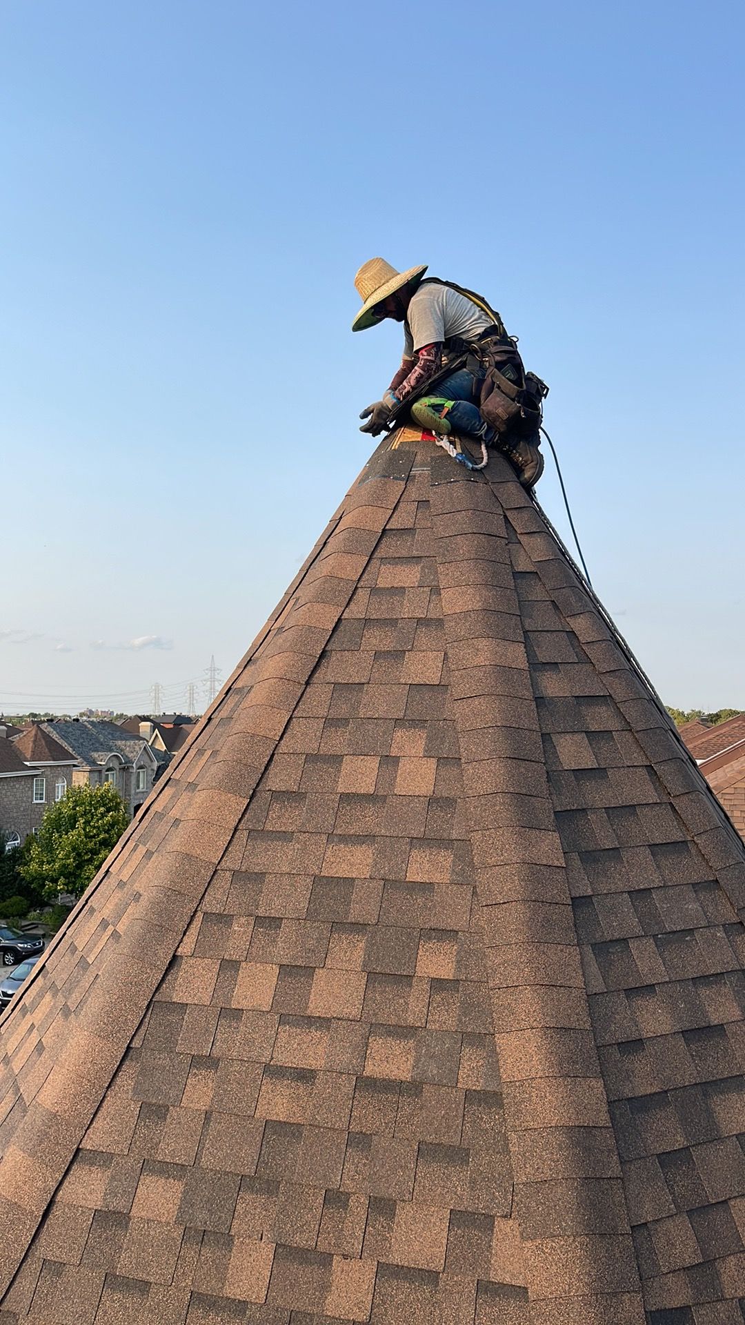 Un couvreur coiffé d'un chapeau de paille est assis au sommet d'un haut toit de bardeaux bruns, sous un ciel d'un bleu limpide.