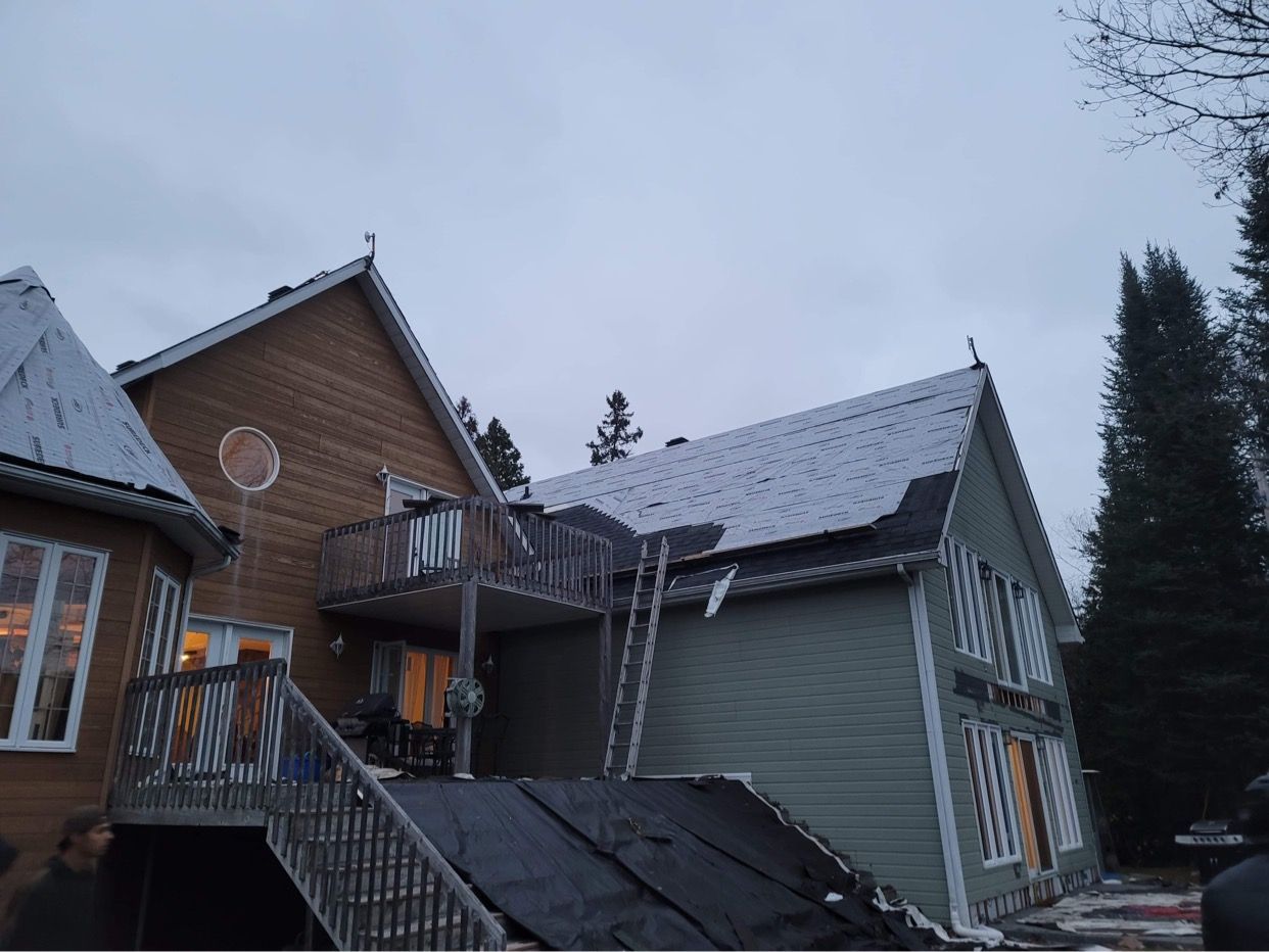Une maison à deux étages en cours de remplacement de toiture, avec une sous-couche noire partiellement installée et une échelle.