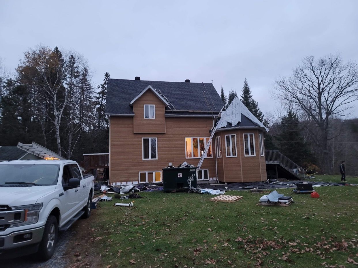 Une maison beige à deux étages en cours de rénovation, avec une benne à ordures, une échelle et une camionnette blanche dans la cour.