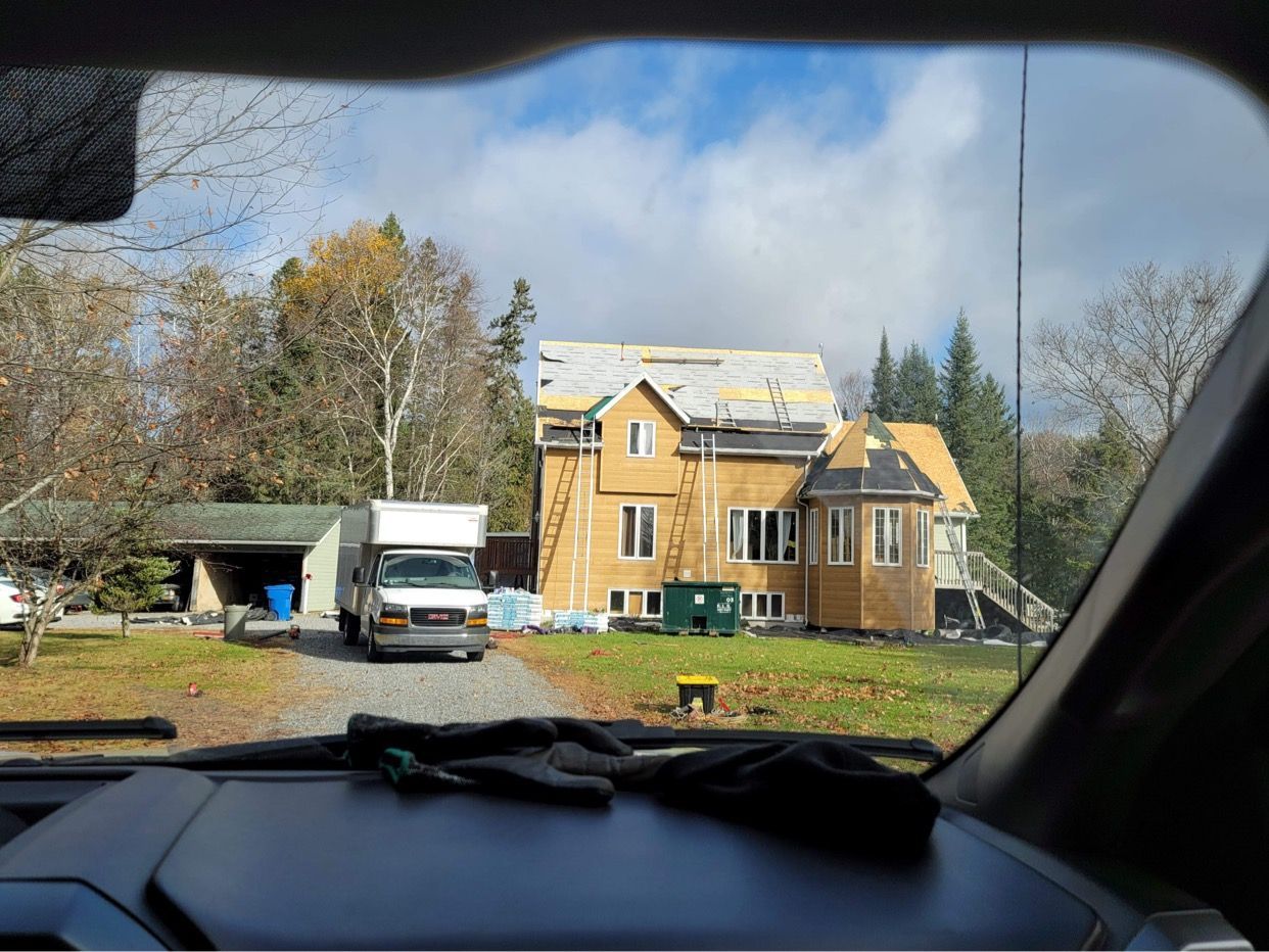 Une maison à deux étages en construction, avec un camion blanc garé dans l'allée, vue depuis l'intérieur d'un véhicule.