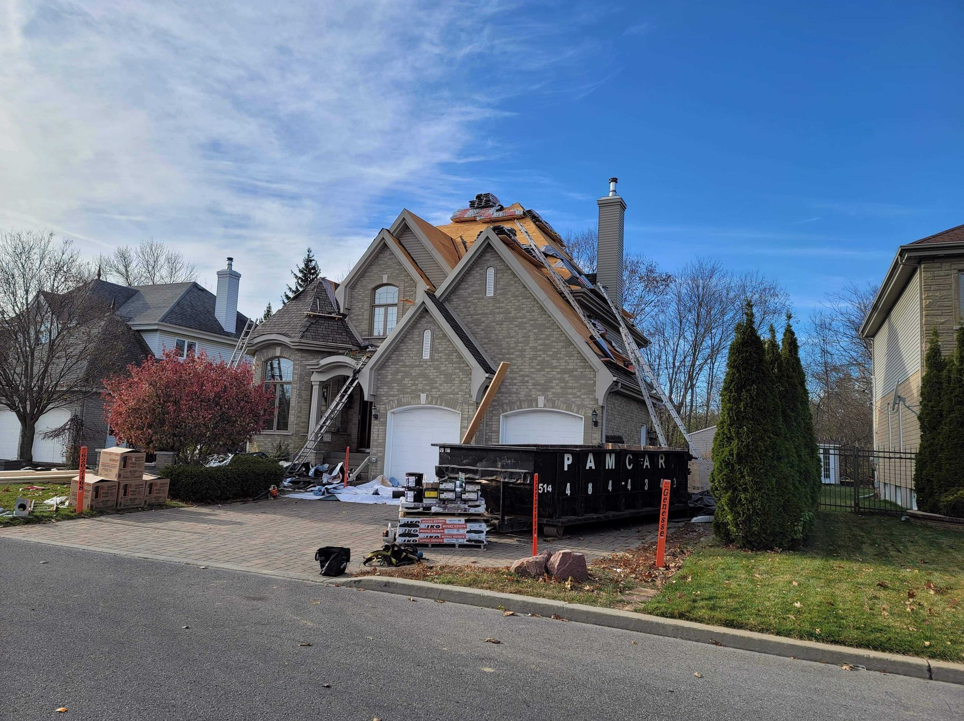 Une maison en pierre en cours de réparation de toiture, avec sa charpente en bois apparente, une benne à ordures dans l'allée et un ciel bleu dégagé.