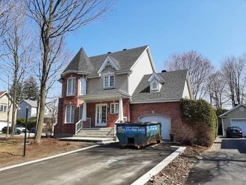 Maison de deux étages en briques et bardage, avec une tourelle et une allée où se trouve un grand conteneur à déchets bleu.