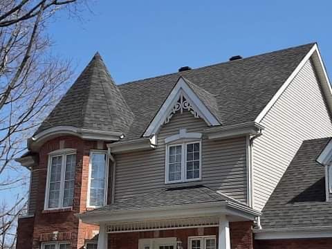 L'extérieur d'une maison à plusieurs étages présente une tourelle en briques, un bardage clair, des bardeaux d'asphalte gris et une garniture de pignon ornée.