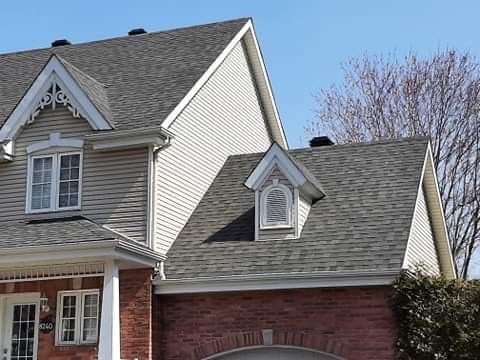 Une maison à deux étages avec un revêtement extérieur beige, des niveaux inférieurs en briques, des toits en bardeaux gris et deux lucarnes à pignon.