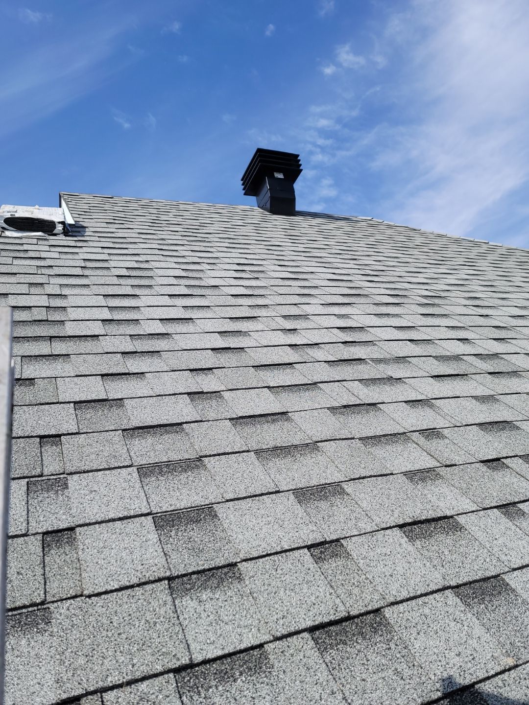 Un toit pentu en bardeaux d'asphalte gris sous un ciel d'un bleu éclatant, avec une cheminée noire près du faîte.