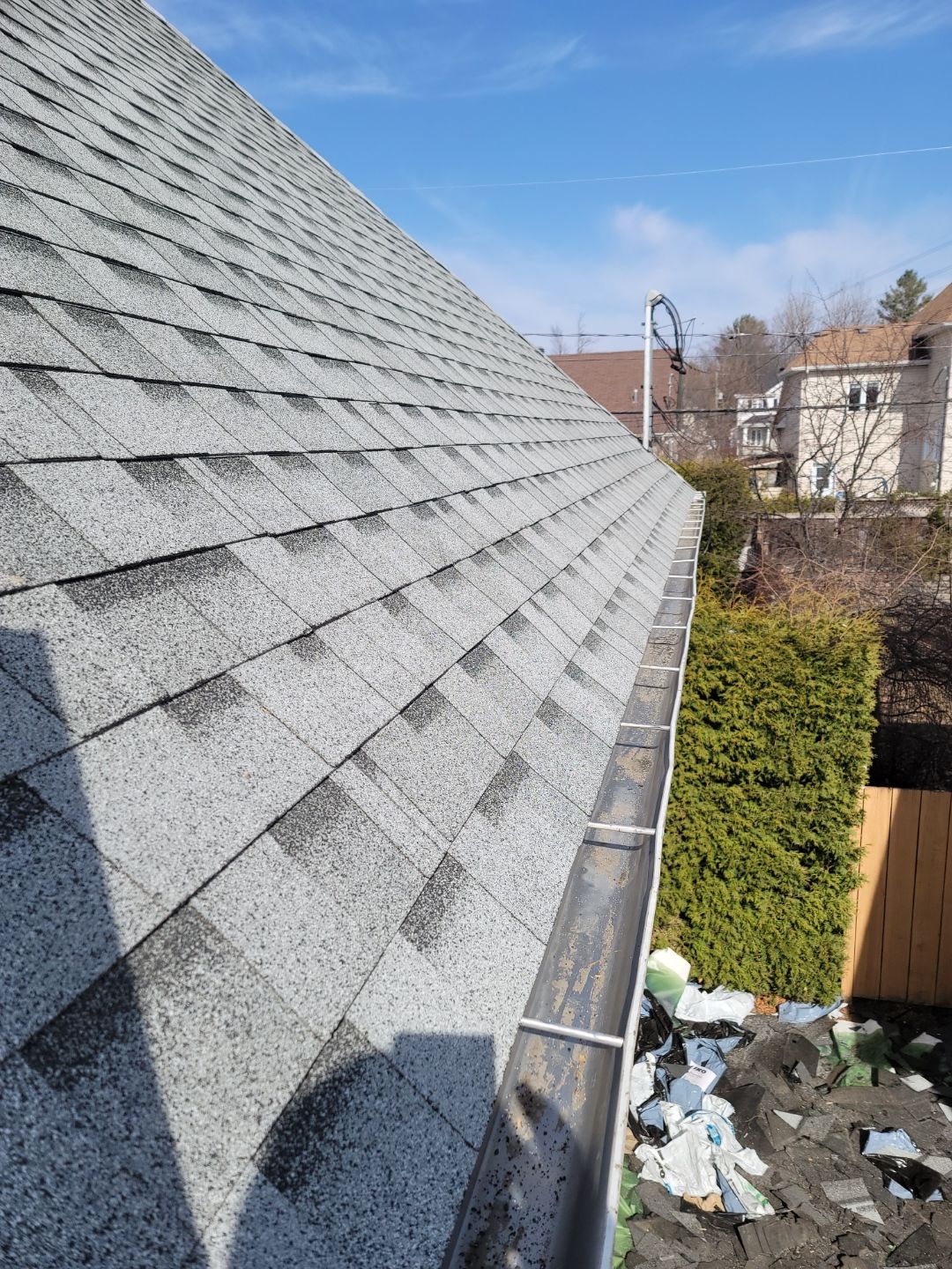 Vue plongeante le long du bord d'un toit en bardeaux vers une gouttière métallique, avec un quartier résidentiel en arrière-plan.