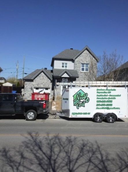 Un camion noir tractant une remorque de travail blanche avec un logo vert est garé devant une maison en pierre grise à plusieurs étages.