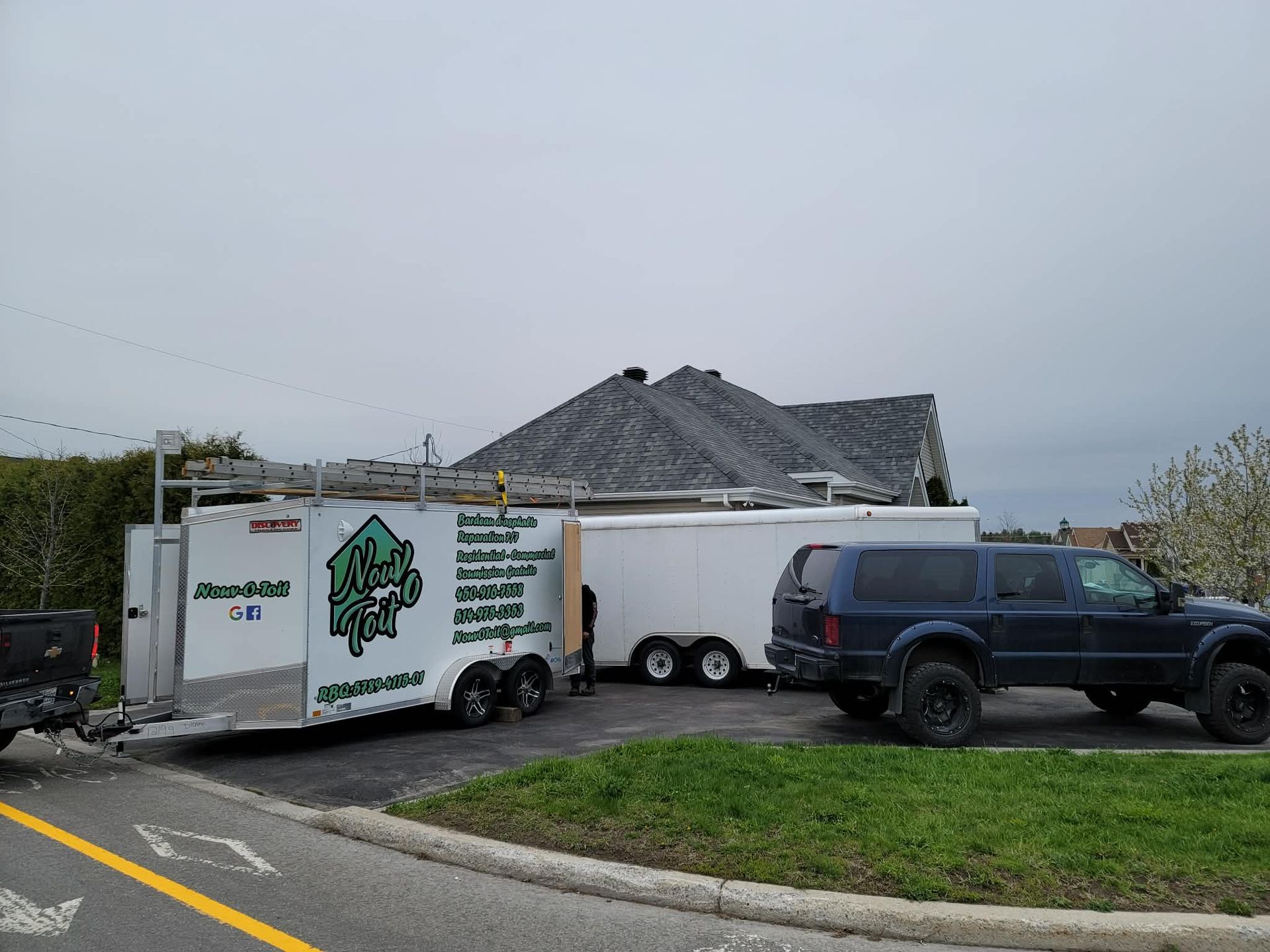 Un camion de service et deux remorques blanches sont stationnés sur le trottoir devant une maison dont la toiture est en cours de remplacement.