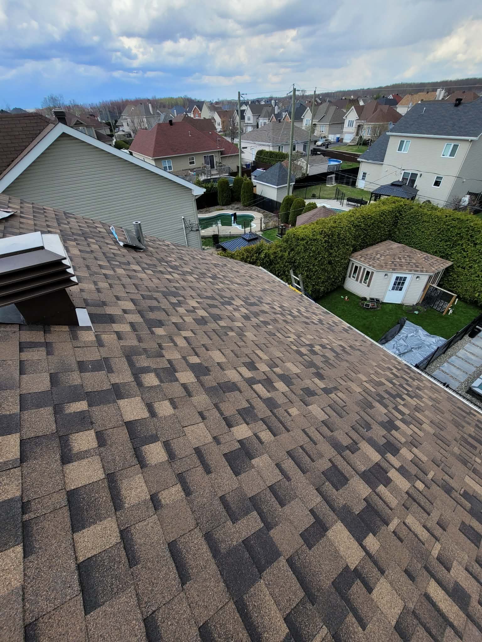 Vue depuis un toit en bardeaux d'asphalte brun surplombant un quartier résidentiel avec des maisons, des jardins et des arbres.