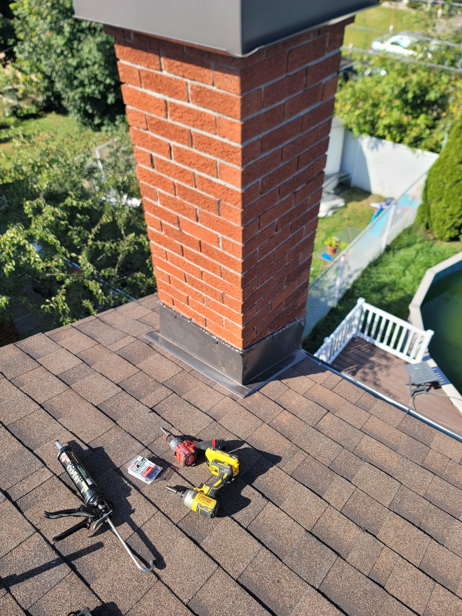 A brick chimney on a shingled roof with a caulk gun, drill, and small hardware parts laid out on the shingles.