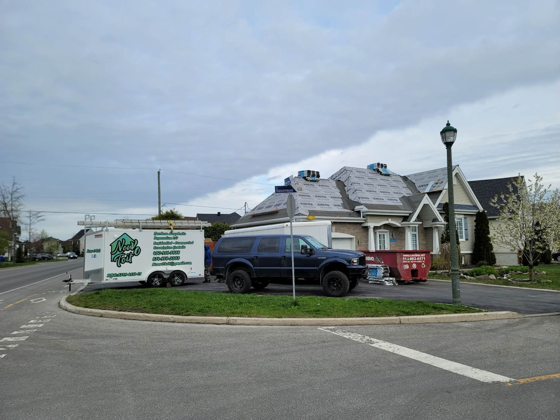 Un SUV foncé et une remorque de chantier blanche étaient garés devant une maison en cours de réparation de toiture par temps nuageux.