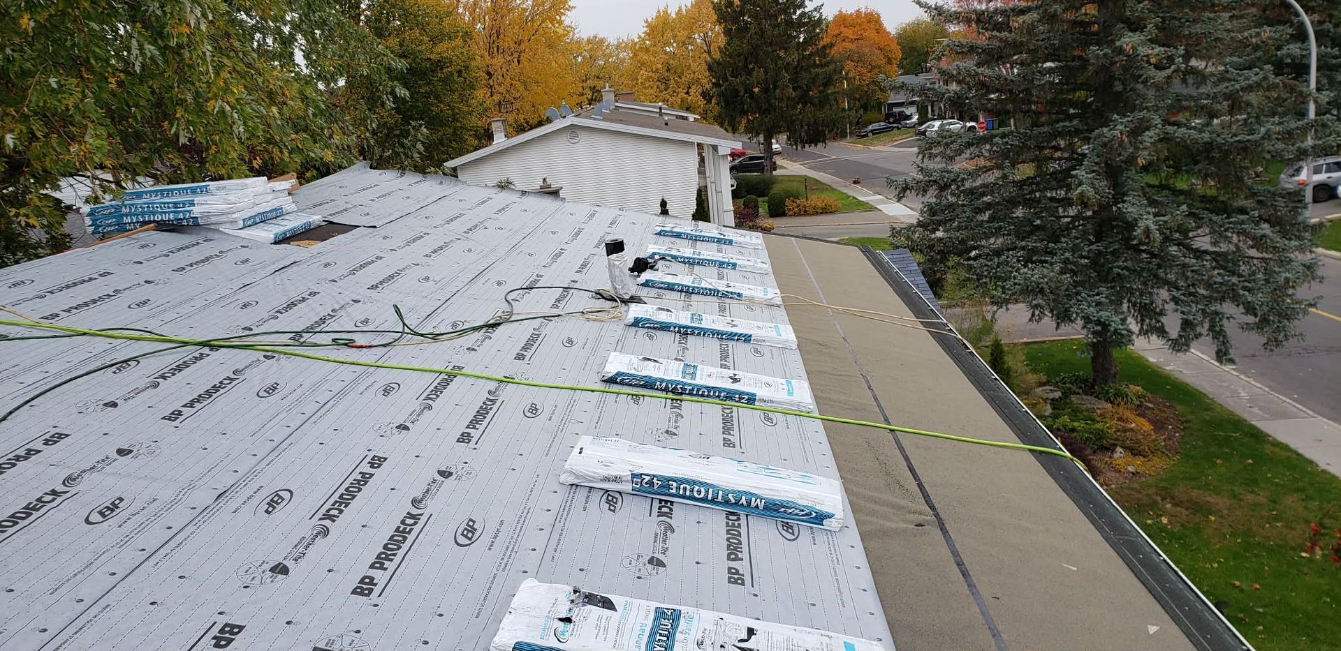 Un ouvrier du bâtiment se tient sur un toit recouvert d'une sous-couche grise aux joints étanches, surplombant une rue du quartier.