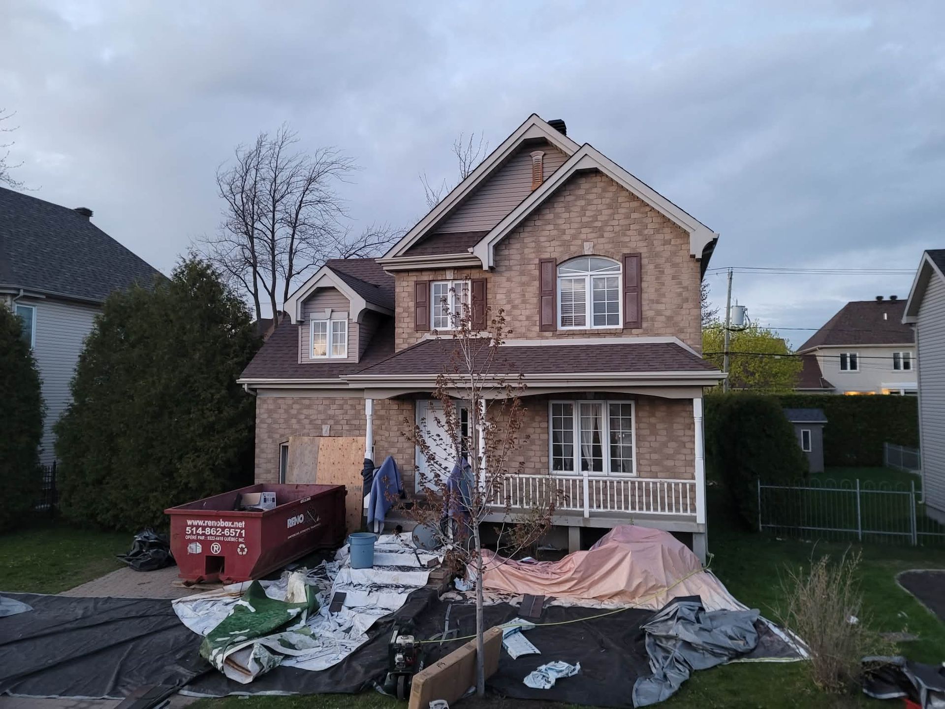 Une maison en briques de deux étages en cours de rénovation, avec un conteneur à déchets dans l'allée et des bâches de protection sur la pelouse.