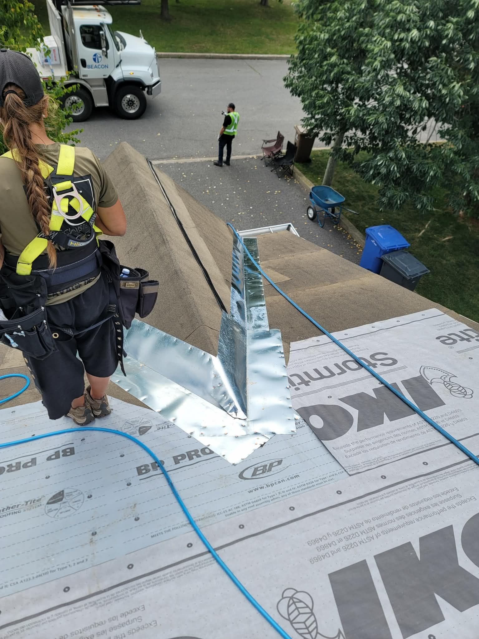 Un couvreur se tient debout sur un toit en pente, portant un harnais de sécurité ; un ouvrier et un camion sont visibles au sol en contrebas.