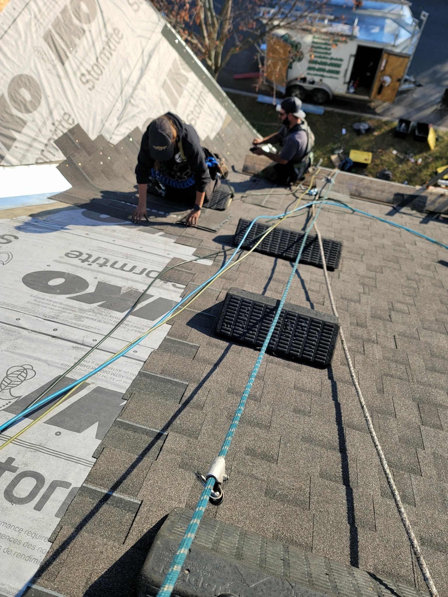 Two workers in safety harnesses install shingles on a steep, sunlit residential roof with protective rope systems.