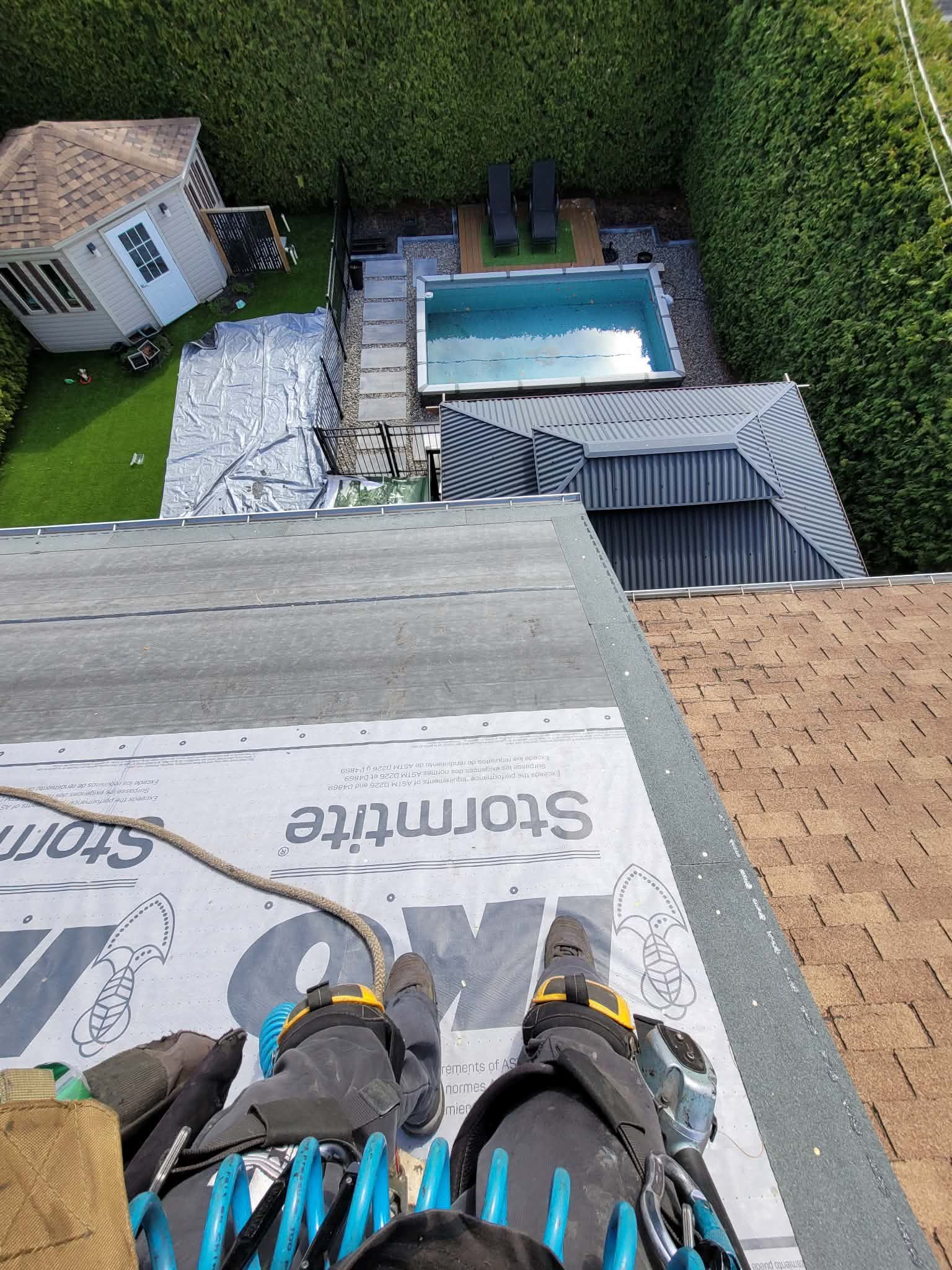 Vue depuis un toit montrant les pieds d'un travailleur et son harnais de sécurité, donnant sur une cour arrière avec piscine, gazebo et abri de jardin.