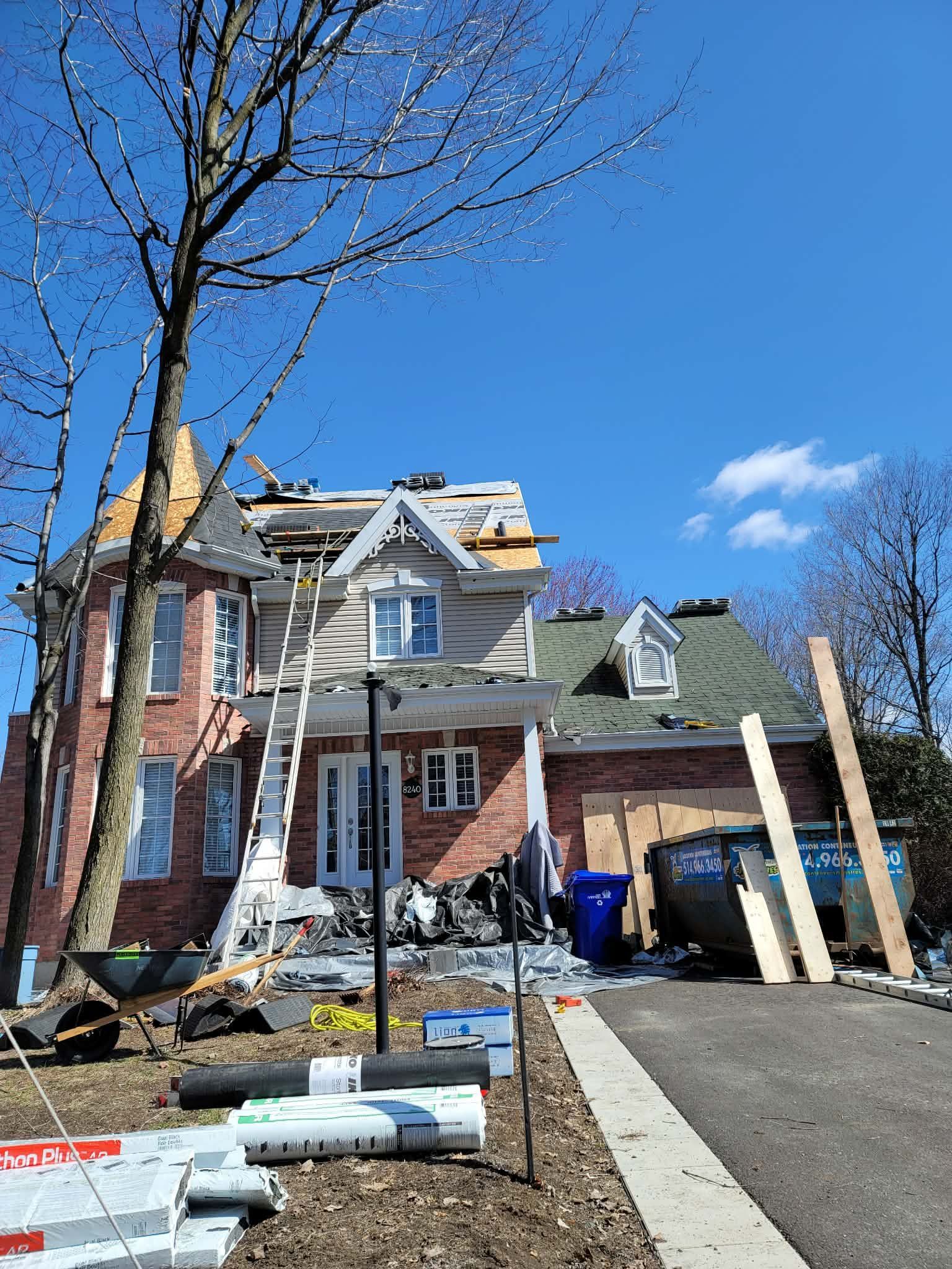 Une maison en briques rouges, dont le toit est partiellement démonté et en cours de réparation, avec une échelle et un conteneur à ordures dans l'allée.
