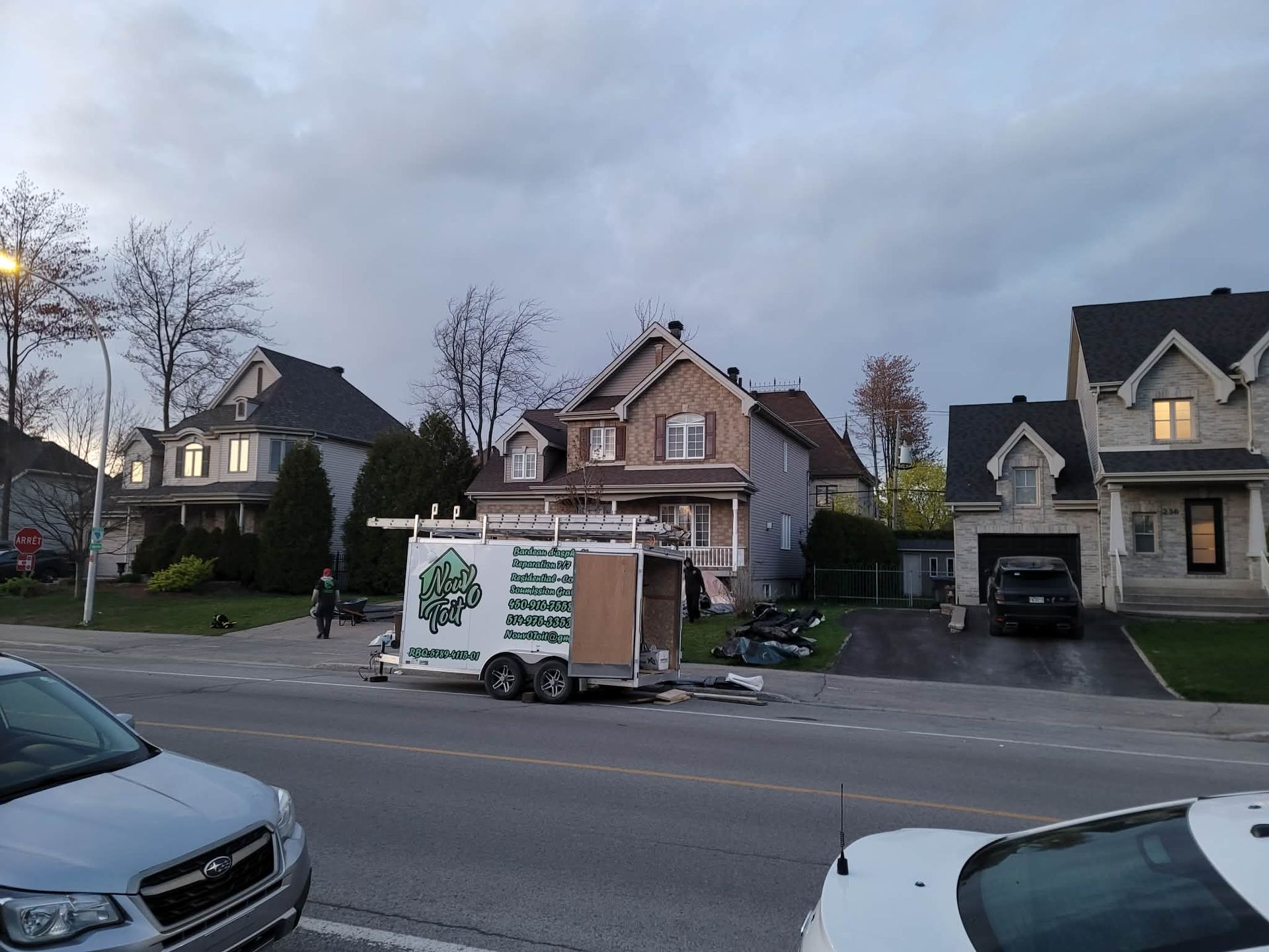 Une remorque utilitaire blanche garée dans une rue de banlieue, devant des maisons résidentielles à deux étages, au crépuscule.