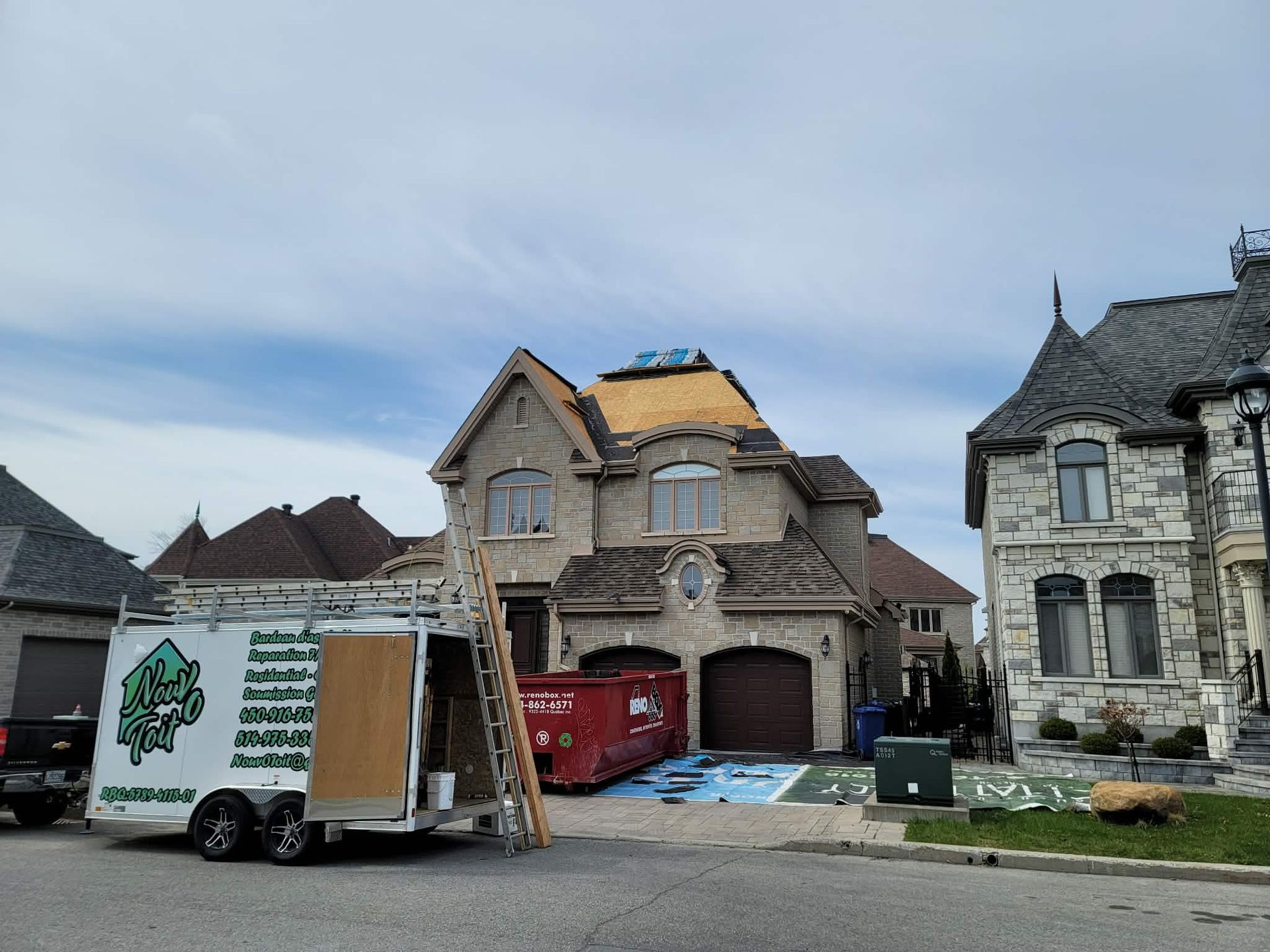 Une maison d'habitation en cours de réparation de toiture, avec une remorque garée devant et un grand conteneur à ordures rouge dans l'allée.