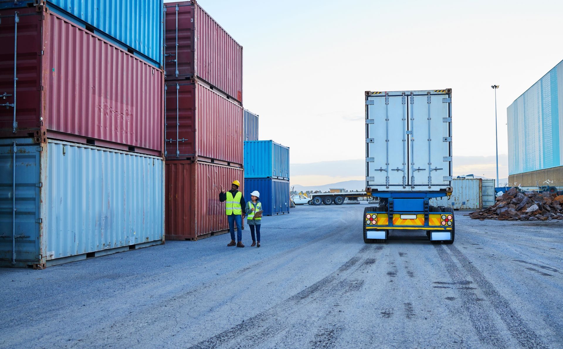 Two workers in safety vests walking by shipping containers near a truck trailer.