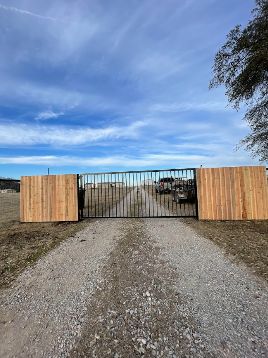 Gravel driveway leading to a closed black metal gate flanked by wooden fences under a blue sky.