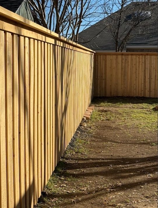 Wooden fence encloses a backyard with brown dirt and patches of green grass under a clear blue sky.