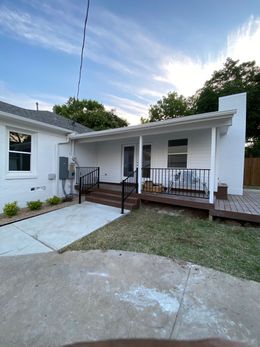 White brick house with a dark brown deck and railing, steps leading to a covered porch under a blue sky.