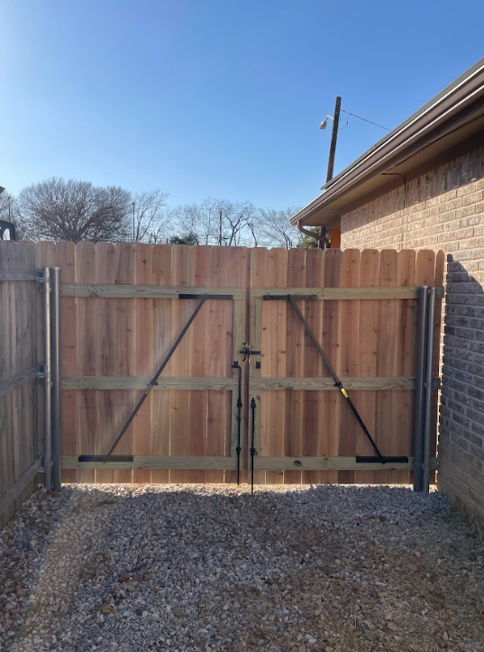 Wooden double gate in a gravel driveway, attached to a brick building and wooden fence, under a blue sky.