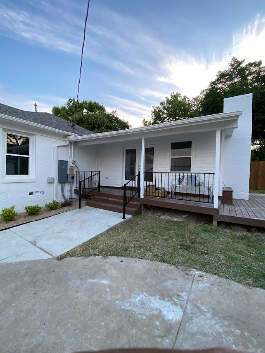White house with covered porch, brown deck, and black railing. Concrete walkway in front.