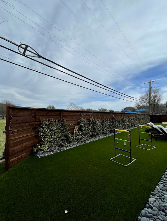 Wooden fence with greenery along it, artificial turf, and a cloudy sky.