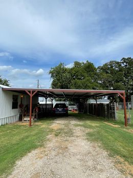 Gravel driveway leads to a car under a carport. Green grass surrounds. Cloudy blue sky overhead.