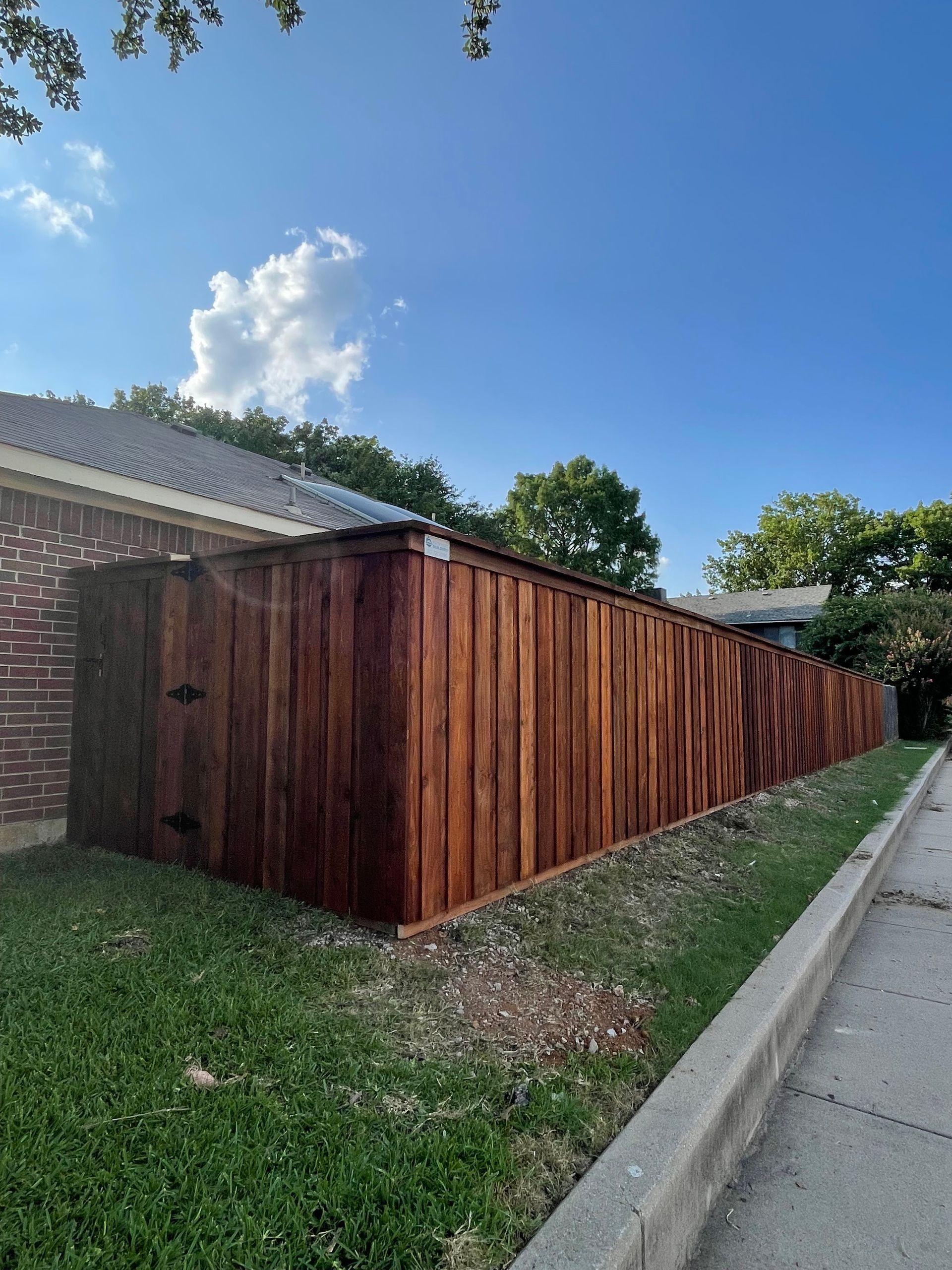 Brown wooden fence next to a grassy lawn and sidewalk under a blue sky.