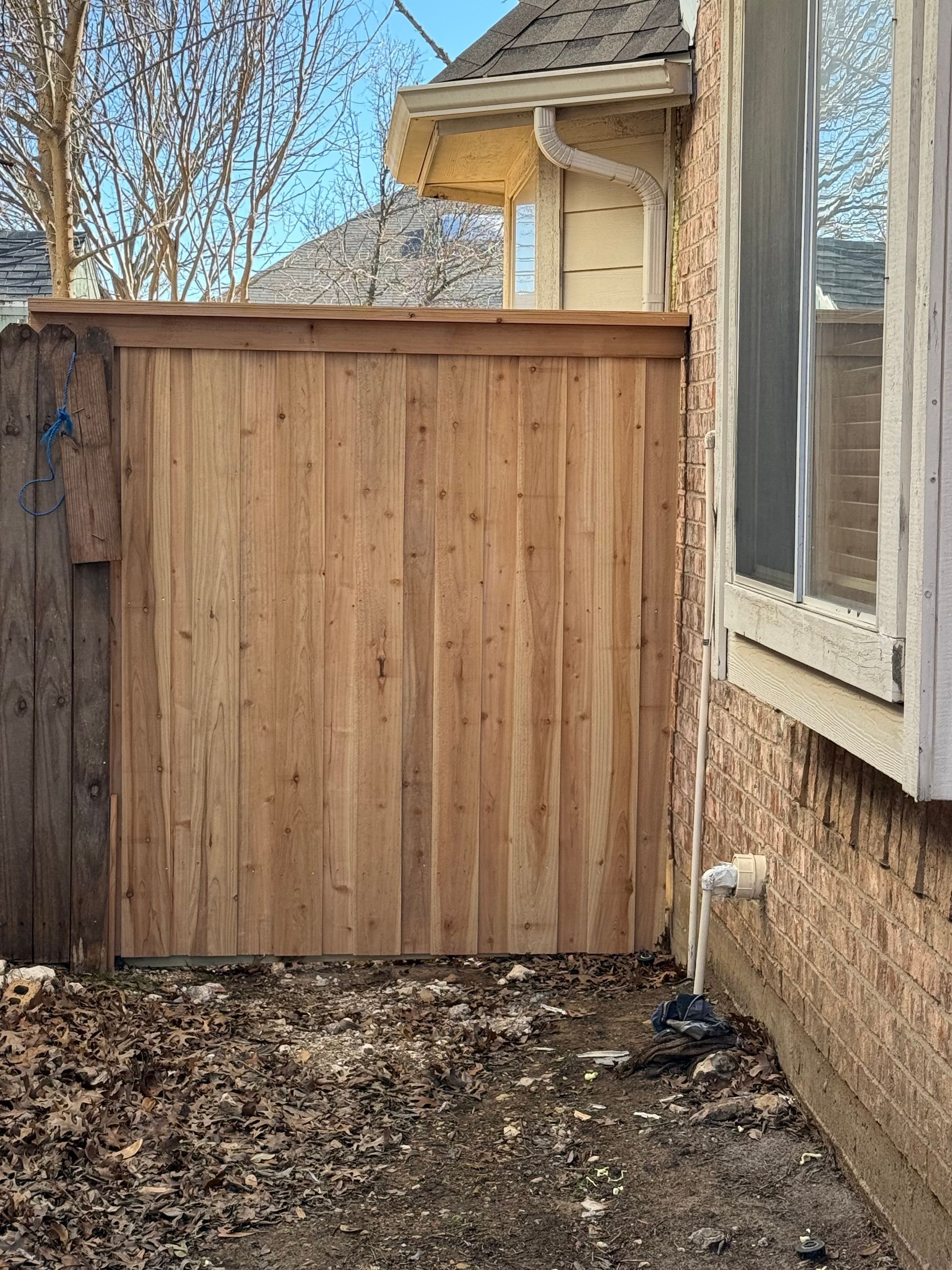 Wooden fence section next to a brick wall with a window. Dry dirt foreground, clear sky.