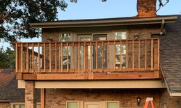 A second-story wooden deck with railings extends from a brick house, featuring glass doors and a chimney.
