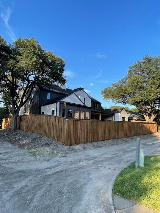 Modern house behind a wooden fence, with black and white exterior under a blue sky, trees on each side.