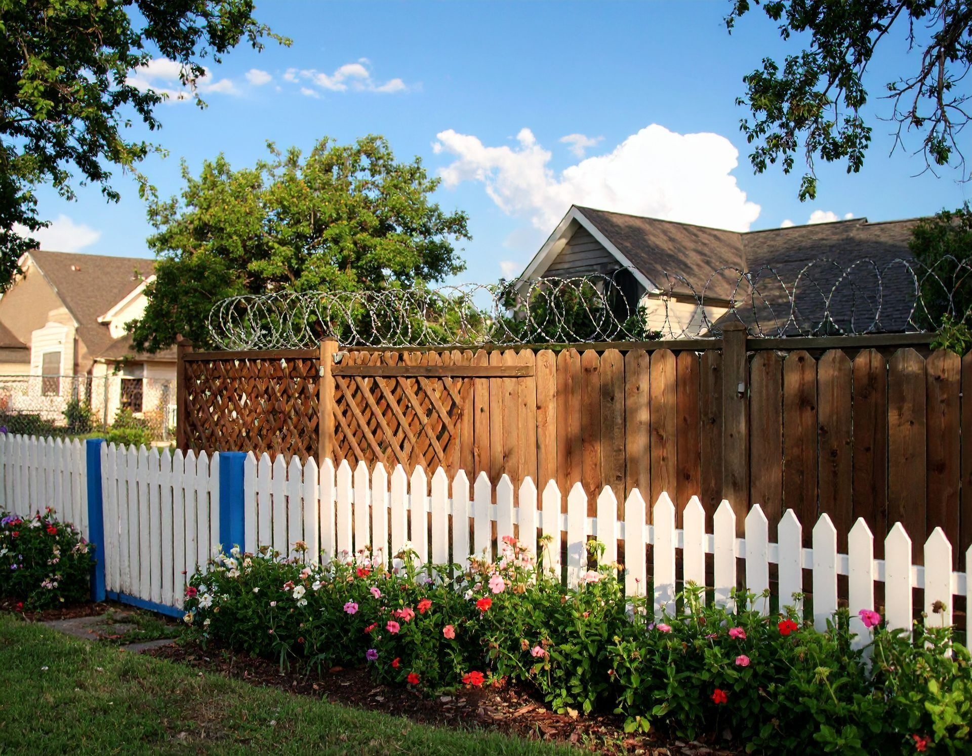 White picket fence with flowers, leading to a wooden fence topped with razor wire, houses in the background.