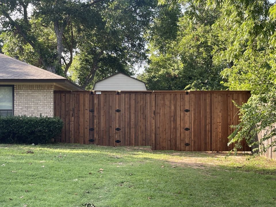 A brown wooden privacy fence in front of a house, with green grass and trees in the background.