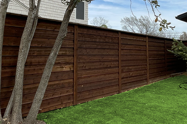 A dark brown wooden fence in a yard with green grass and a tree trunk in the foreground.
